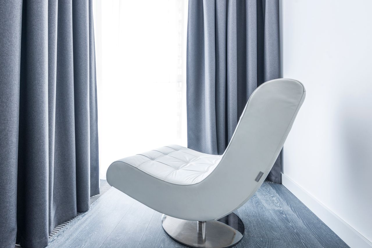A modern white leather lounge chair in a minimalist room with grey drapes and natural lighting.